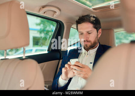 Young businessman sending a text message on a cellphone while sitting in the backseat of a car driving through the city Stock Photo