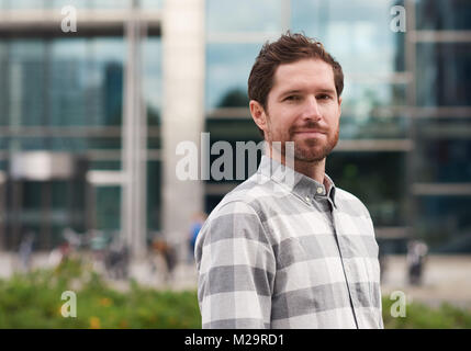 Trendy dressed bearded man smiling while standing alone in front of buildings on a street in the city Stock Photo