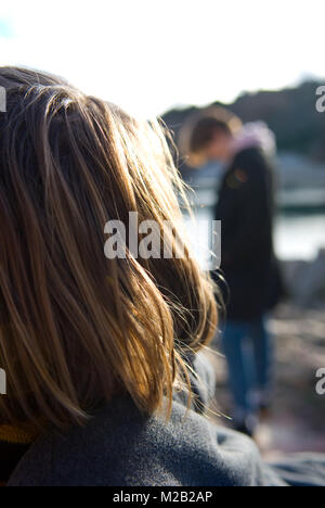 macro detail shot of woman’s hair in sharp focus in foreground with blurred teenager looking down at ground in a dejected manner Stock Photo