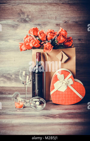 Red wine bottle, two glasses of wine, roses in a paper bag and gift  heart  on wooden table. Valentines day celebration concept. Copy space. Stock Photo