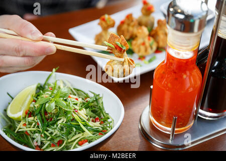 Hand holding one fried wonton with sticks, with vegatable salad and set of sauses standing on a table Stock Photo
