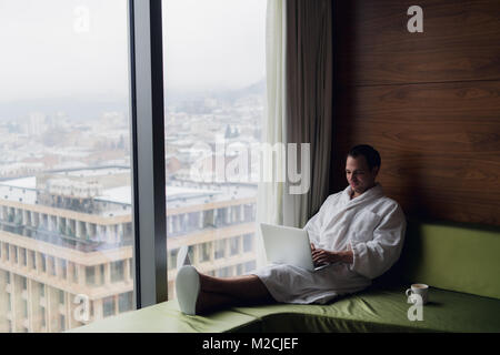 Businessperson working at home or in trip. Side view portrait of handsome young businessman sitting on bed and using laptop. Panoramic window with beautiful dawn city scenery on the background Stock Photo