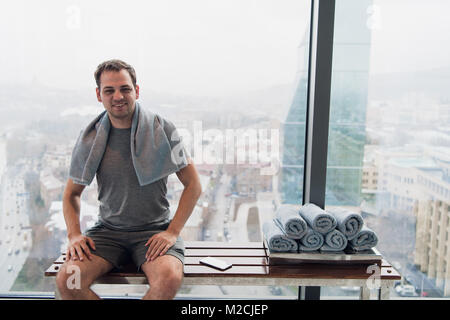Young man in gym after workout, sitting on a bench in front of wide windows overlooking modern city skyscrapers Stock Photo