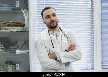Serious male doctor is holding his stethoscope and standing with arms folded in his office looking at view with menace. Stock Photo