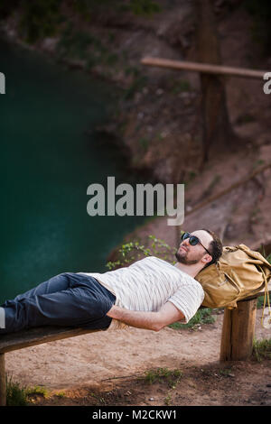 Man Relaxing Outside. Wearing a white T shirt, dark pants, sunglasses, a young guy is lying down on wooden bench against lake in a forest outdoors Stock Photo