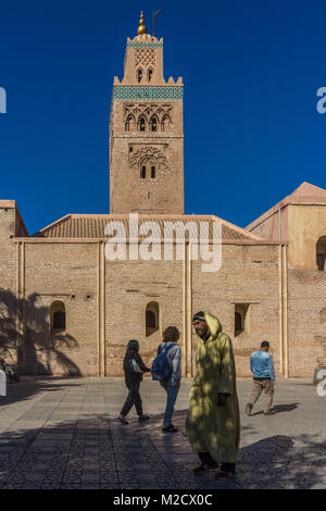 A man wearing a yellow djellaba walks by Koutoubia Mosque and Minaret, in Marrakesh, Morocco, while a group of young tourists walk the other way. Stock Photo