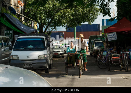 A Burmese couple, possibly husband and wife, with the man driving a cycle rickshaw in  the busy Yangon traffic and the wife sitting passenger. Myanmar Stock Photo