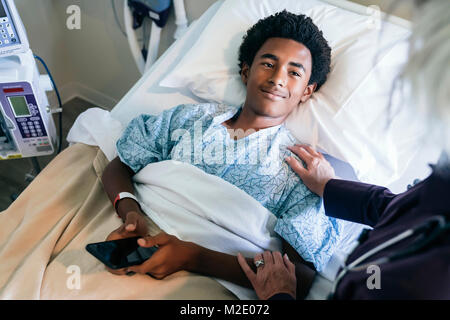 Doctor comforting boy laying in hospital bed holding cell phone Stock Photo