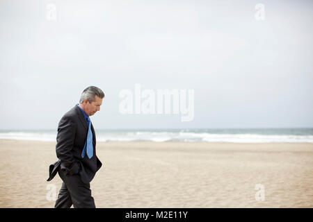 Pensive Hispanic businessman walking on beach Stock Photo