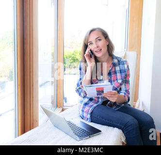 Caucasian woman holding paperwork and talking on cell phone near window Stock Photo