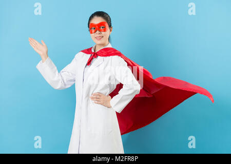 confidence pretty woman clinic doctor wearing superhero clothing face to camera and making introduce gesture isolated on blue background. Stock Photo
