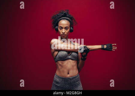 Fit young woman in sportswear and headphones stretching her arms. Portrait of african woman doing warm up exercises against red background. Stock Photo