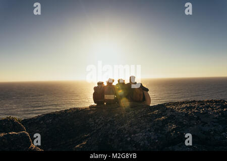 Rear view of friends sitting together on mountain top and enjoying the sunset over sea. Men and women relaxing on mountain top during sunset. Stock Photo