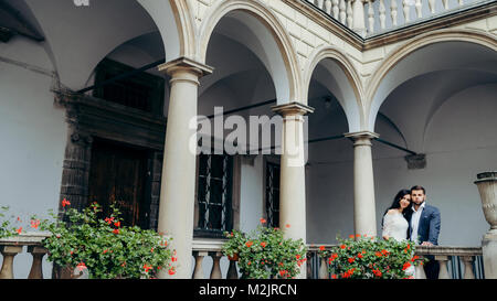 Beautiful newlyweds are enjoying the view while standing on the old balcony decorated with red flowers. The gorgeous bride is leaning on the groom shoulder. Stock Photo