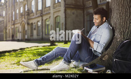 Mixed-race young guy sitting under tree, taking notes in notebook, inspiration Stock Photo