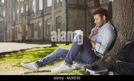 Multiracial young guy sitting with notebook under tree, thinking what to write Stock Photo