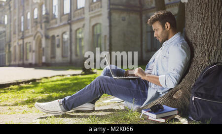 Multinational young man reading information on laptop under tree, concentrated Stock Photo