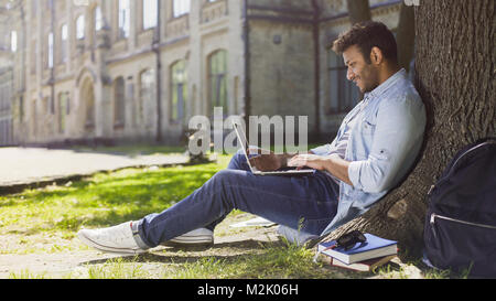 Young mixed-race guy using laptop under tree, smiling at good news, emotions Stock Photo