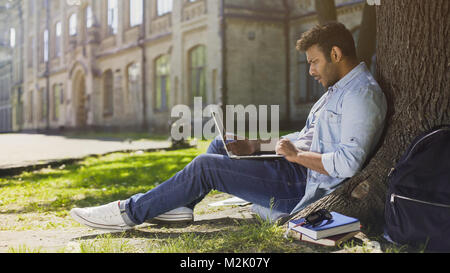 University student under tree, using laptop looking surprised, upsetting news Stock Photo