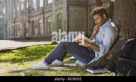 University student sitting under tree reading book with gripping plot, engrossed Stock Photo