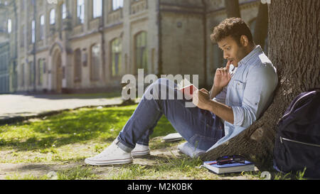 Multiracial young guy sitting under tree, reading interesting book, bookworm Stock Photo