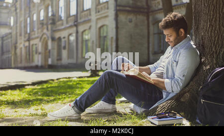 Mixed-race guy sitting under tree in park, reading book, leisure time, hobby Stock Photo