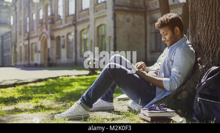 Young man sitting under tree in park, using mobile phone and smiling, good news  Stock Photo