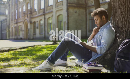 Male sitting under tree, using mobile phone, reacting to newsfeed, confusion Stock Photo