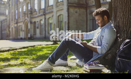 Multinational young man sitting under tree looking at mobile phone screen, smile Stock Photo