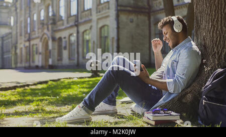 Guy in headphones sitting on grass and leaning against tree, singing to music Stock Photo