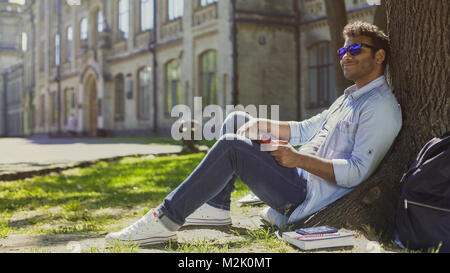 Multiracial man sitting under tree wearing sunglasses, positive mood, energy Stock Photo