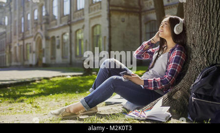 Female student sitting on grass, leaning against tree, listening to music, relax Stock Photo