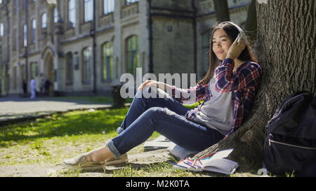 Multinational young woman in headphones sitting under tree, listening to music Stock Photo