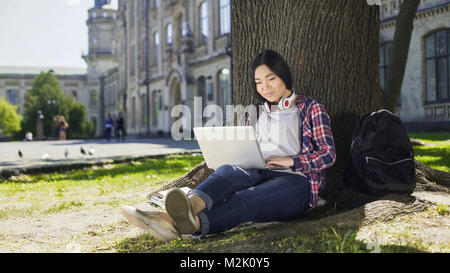 Young university student using laptop with smile on face, sitting under tree Stock Photo