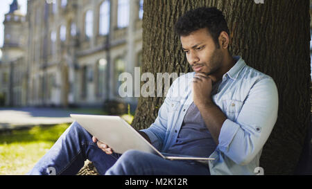 Mixed-race young man sitting under tree, looking at laptop, important letter Stock Photo