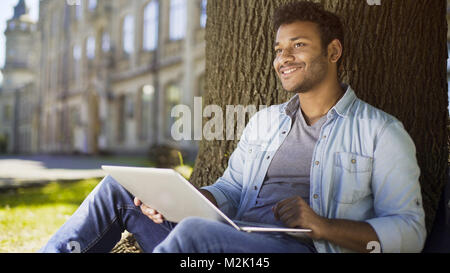 Mixed-race young man with laptop sitting under tree, dating website profile Stock Photo