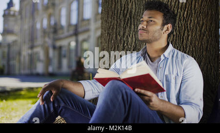 Young man sitting under tree reading book and thinking, fiction literature Stock Photo
