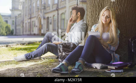 Multiracial guy talking on cellphone under tree, girl taking notes in notebook Stock Photo