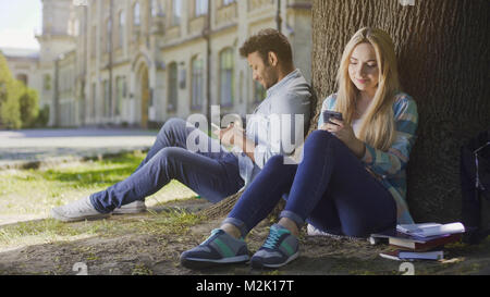 Multiracial guy and girl sitting under tree using mobile phones smiling, happy Stock Photo