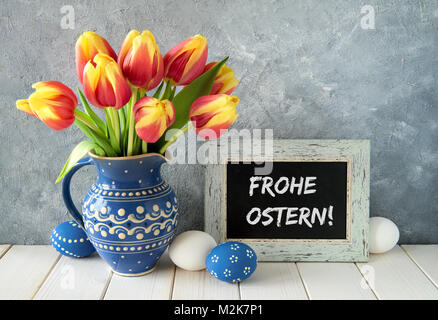 Red-yellow tulips in blue ceramic pitcher with Easter eggs and a blackboard on gray background. Text "Frohe Ostern" means "Happy Easter" in German. Stock Photo