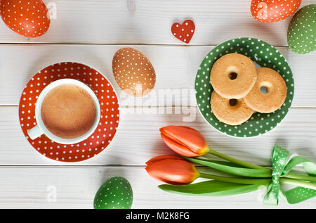 Top view on white wooden table with espresso cup, plate of cookies, Easter eggs and tulips in red and green. Both eggs and plates have polka dot desig Stock Photo