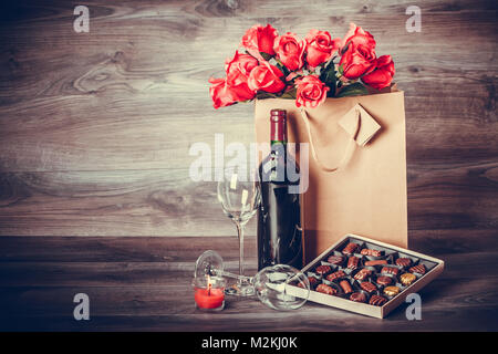 Red wine bottle,two glasses of wine, box of chocolates and roses in a paper bag on wooden table. Valentines day celebration concept. Copy space. Stock Photo