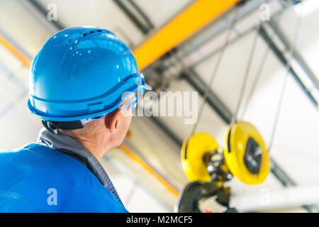 Caucasian Production Line Technician in Blue Hard Hat Checking on the Overhead Warehouse Crane Installed Right Above His Head. Heavy Duty Lift. Head P Stock Photo