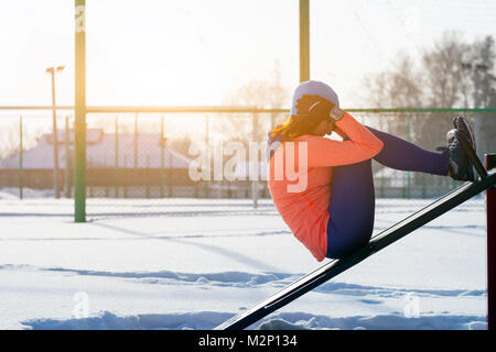 A young woman in a bright blue hat, orange sweatshirt and elk skewers presses a press on a vertical simulator on a sports field on a bright winter day Stock Photo