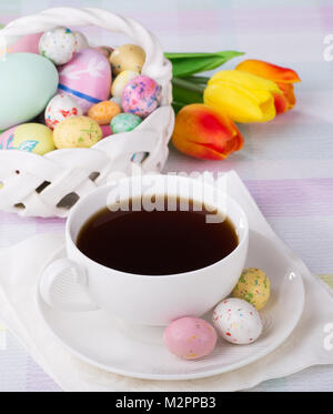 Cup of coffee with Easter candy on saucer with basket of treats and flowers in background Stock Photo