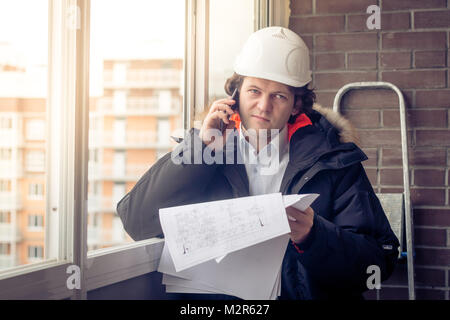 Portrait of concerned unshaven builder telling by mobile while keeping different projects in hand. Communication and work concept. Soft focus, toned Stock Photo