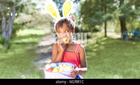 Cute and beautiful African American girl wearing bunny ears, eats with joy Easter marshmallow candy while holding Easter basket full of eggs. Girl sit Stock Photo