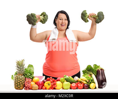 Overweight woman exercising with broccoli dumbbells behind a table with fruit and vegetables isolated on white background Stock Photo