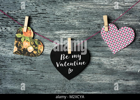 some hearts hung with clothespins in a clothes line and a black heart-shaped signboard with the text be my valentine written in it, against a gray rus Stock Photo