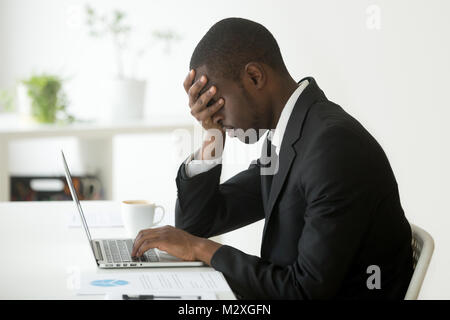 Tired depressed black businessman at workplace frustrated by business failure feeling exhausted covering face with hand, upset sleepy african office w Stock Photo
